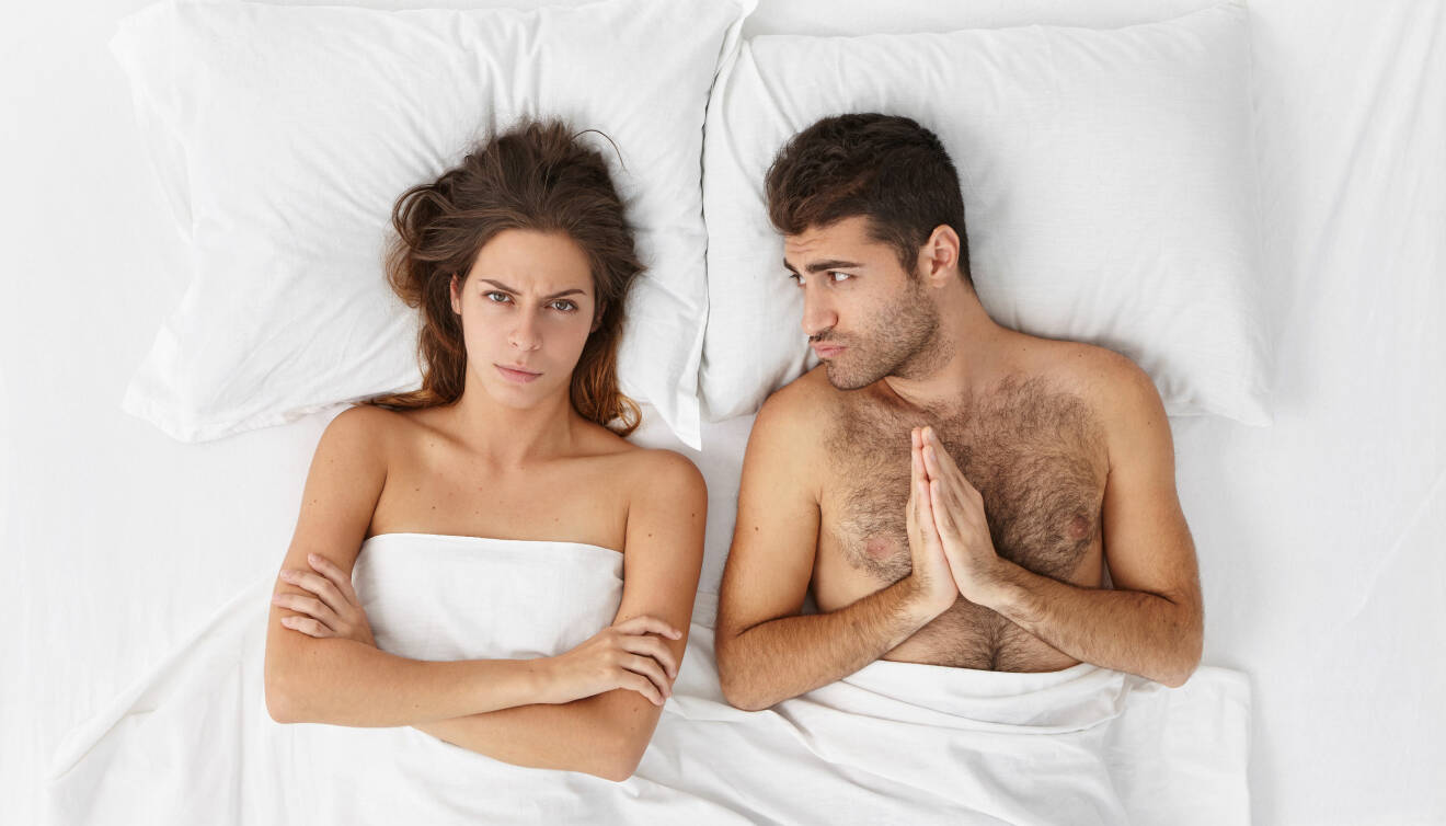 Kvinna och man ligger i en säng. Mannen ser bedjande ut.
