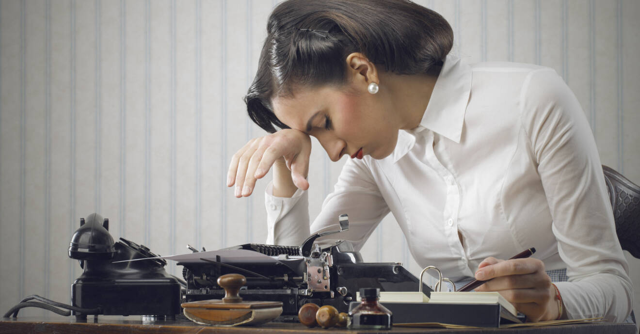 En kvinna i 1950-talsfrisyr ser trött ut vid ett skrivbord.