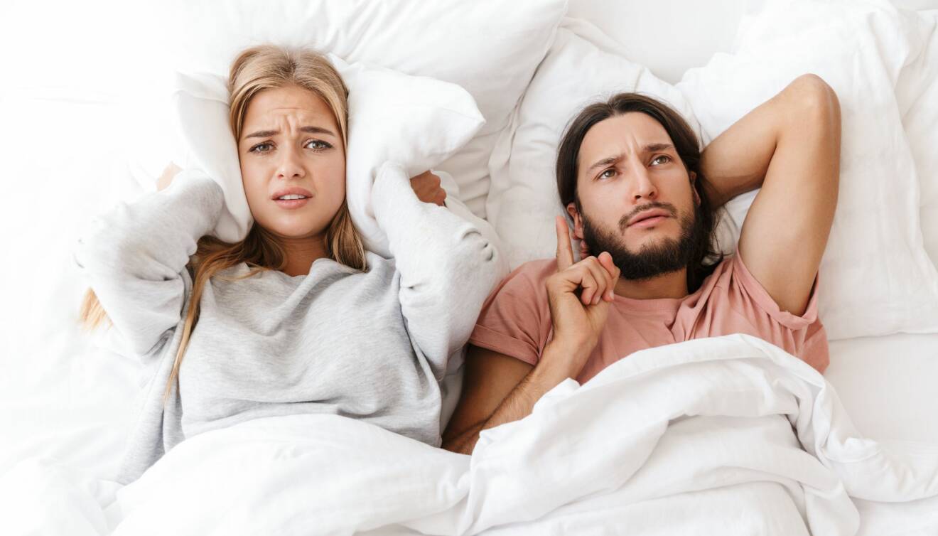 Ett heterosexuellt par ligger bredvid varandra i sängen. Kvinnan håller en kudde för öronen, och mannen argumenterar.