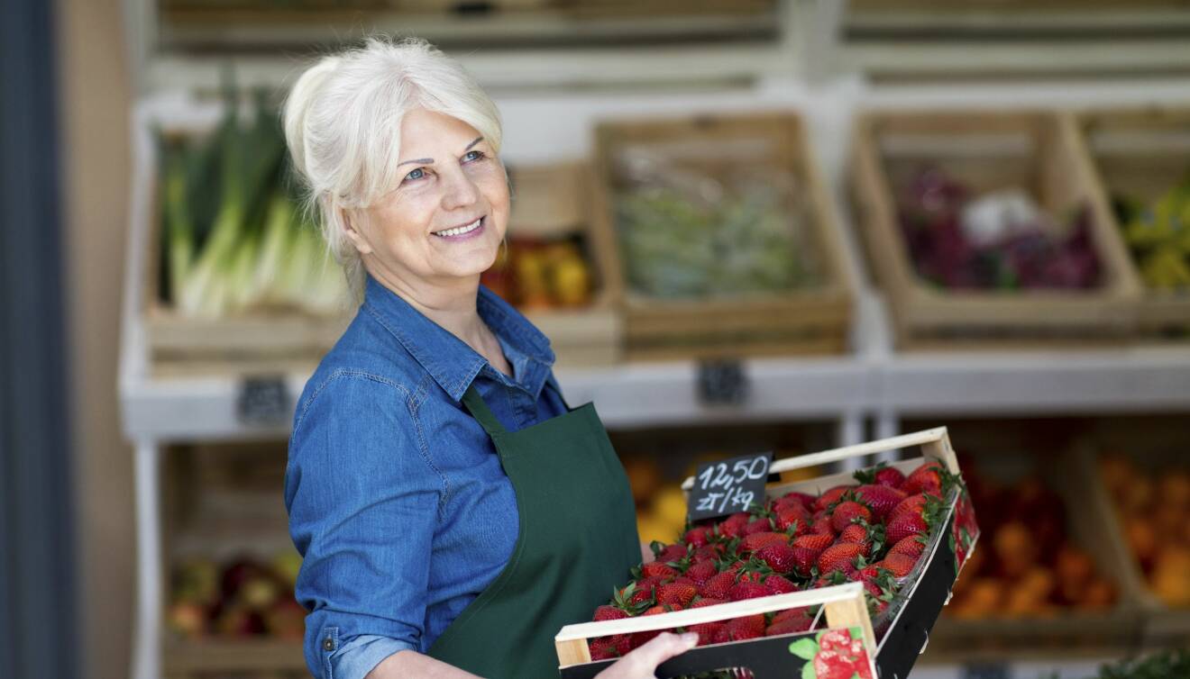 Kvinna i övre medelåldern, butikspersonal, ler glatt medan hon arbetar i grönsaksdisken.
