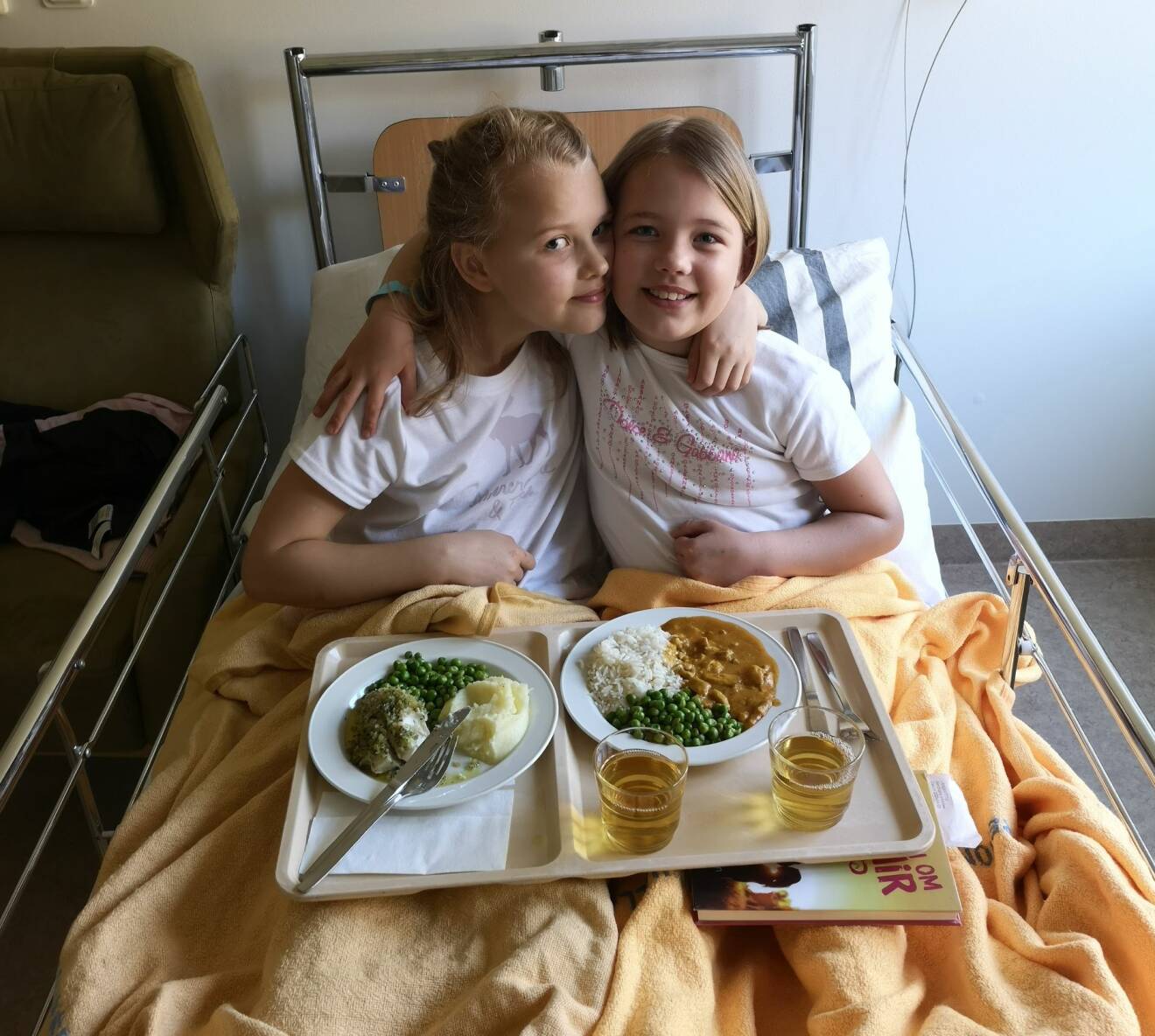 Elsa och Ellen är på sjukhuset för provtagning och sitter tillsammans i en sjukhussäng och har brickor med mat i knät