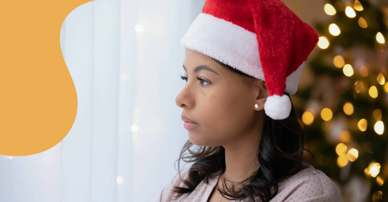 En kvinna i tomteluva tittar dystert ut genom ett fönster. I bakgrunden syns en klädd julgran.