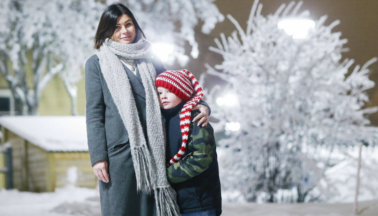 Marie bär rock och lång halsduk, sonen röd och vit-randig mössa och vinterjacka och i bakgrunden syns snöklädda träd och hus med snö på taken.