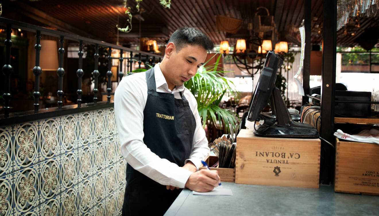 Nas Hosseini står i kassan på restaurang Trattorian i Stockholm och gör anteckningar på ett papper.
