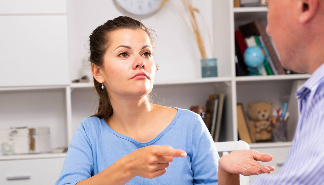 Kvinna i 30-årsålder höjer fingret mot sin man och ser irriterad ut, där de sitter vid köksbordet.