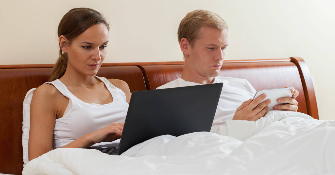 Ung kvinna och ung man i den gemensamma dubbelsängen. Hon är fullt upptagen men sin laptop, och han med sin mobil.