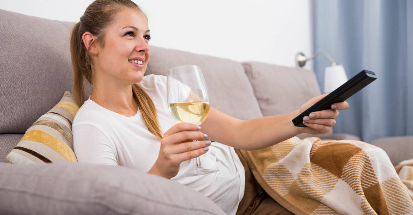 En kvinna liggandes i en soffa med ett glas i ena handen och en tv-dosa i andra.