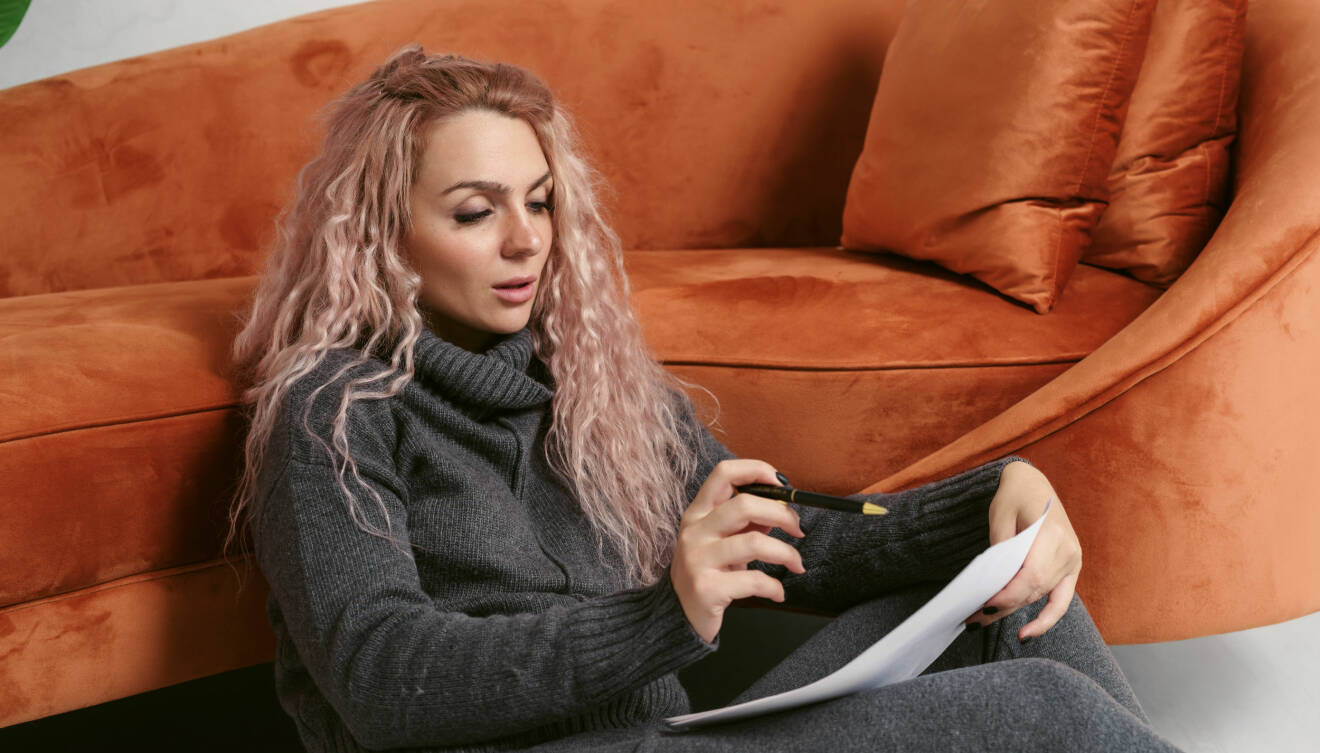 Ung kvinna med långt, blont, lätt rosa-slingat hår sitter på golvet framför kopparfärgad soffa och skriver en lista.