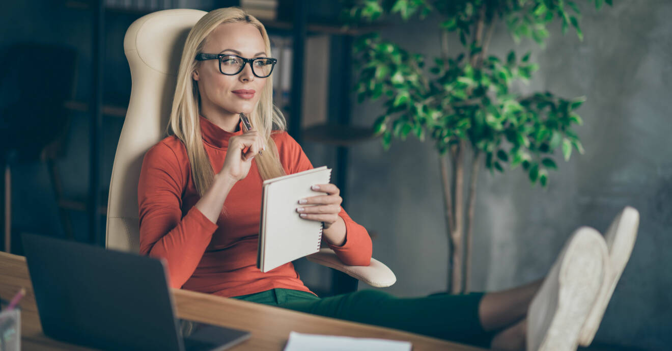 Kvinna i glasögon funderar. Håller ett block i handen vid skrivbord framför laptop.