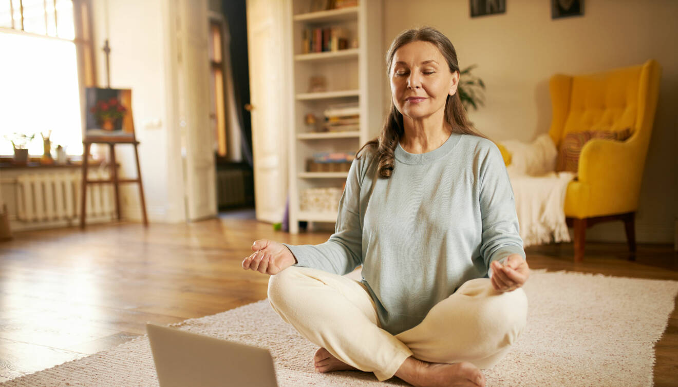 Kvinna mediterar och finner lugn genom att andas framför dator på matta hemma.