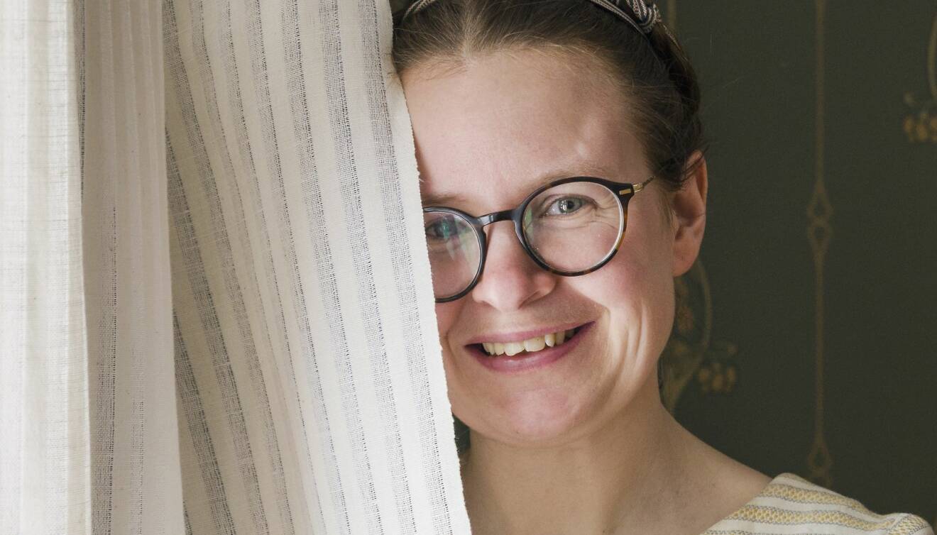 Sara Kånåhols har står framför ett fönster och håller sin egenvävda gardin framför halva ansiktet och berättar om sitt projekt att dokumentera ödehusgardiner.