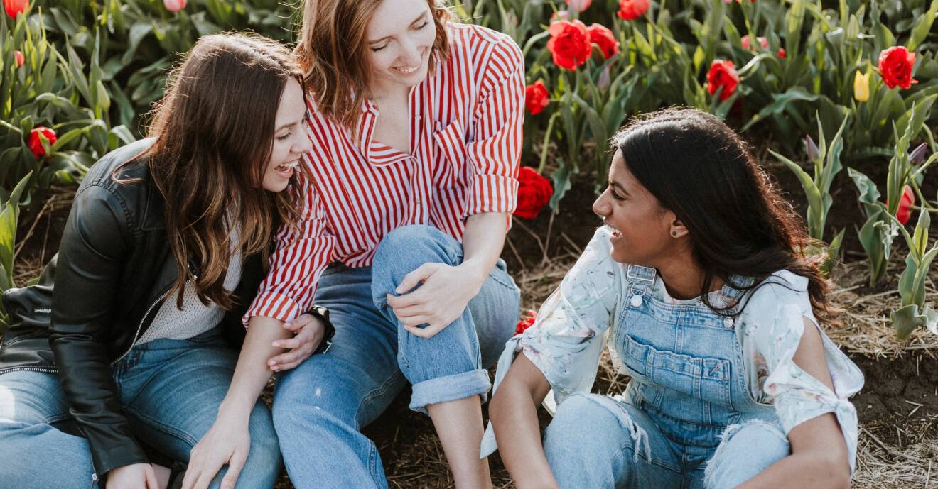 Tre kvinnliga vänner skrattar tillsammans framför röda tulpaner.