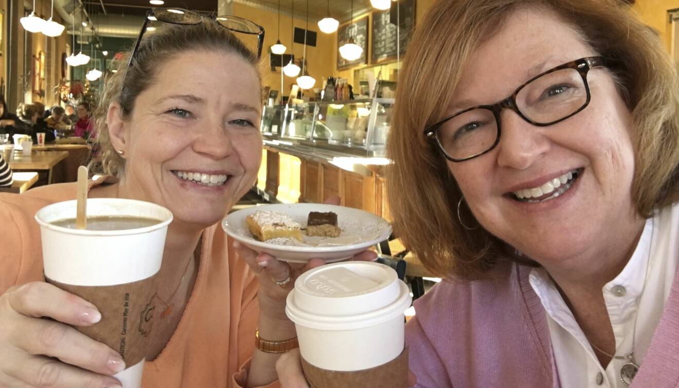 Susan och Michele håller upp kaffemuggar och kakor och ler stort.