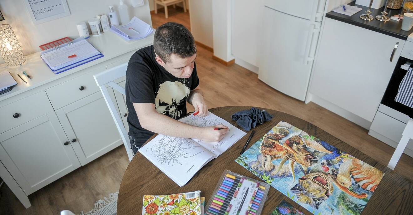 Adam sitter vid köksbordet i sitt kök och färglägger dinosaurier i ett målarblock och bredvid honom ligger ett färdiglagt pussel med dinosauriemotiv.