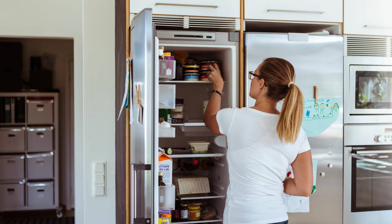 En kvinna står i ett kök och städar ur sin kylskåp. Kvinnan har vit t-shirt och en hästsvans.