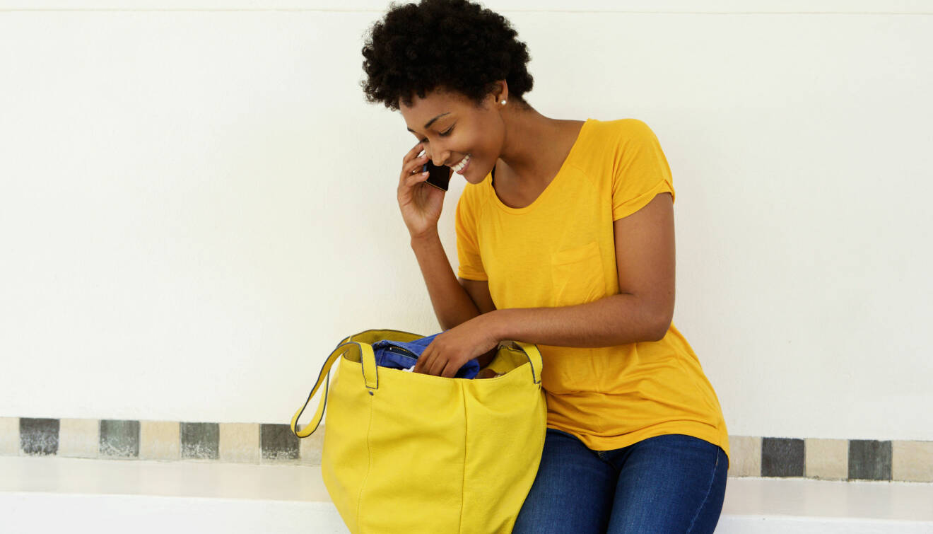 En glad kvinna pratar i mobiltelefon och letar efter något i sin stora gula väska.