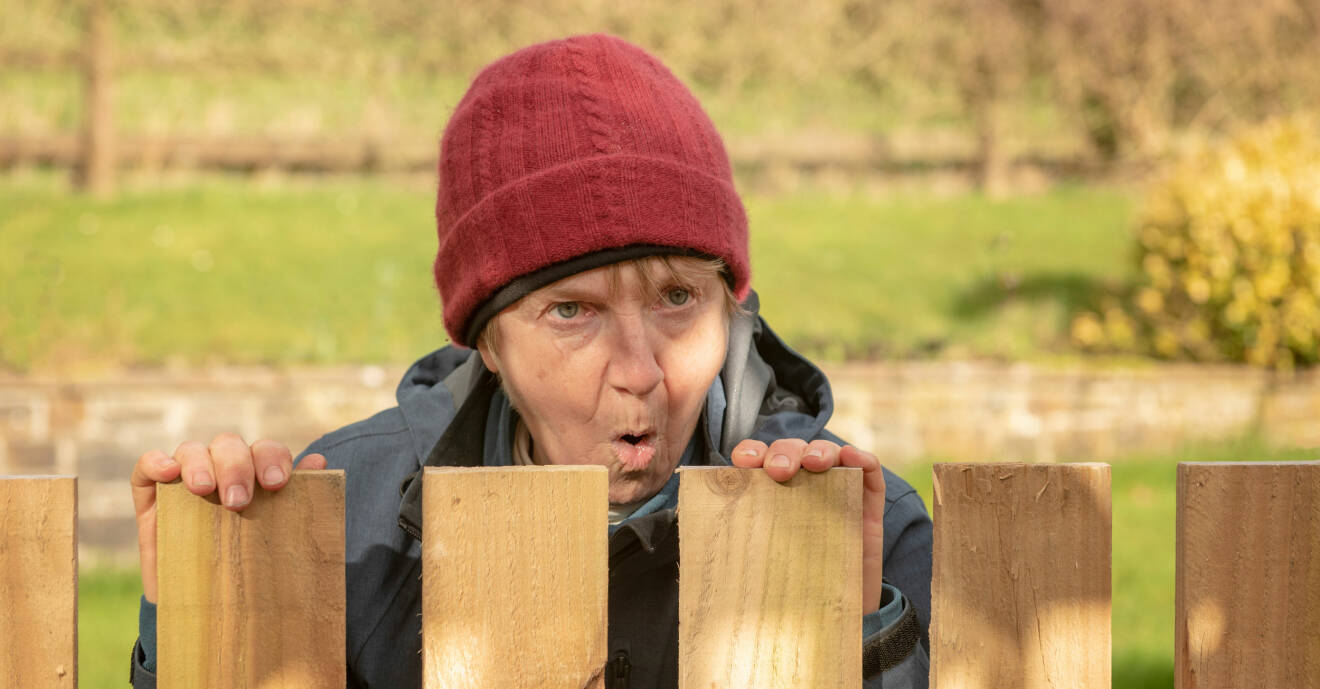 En kvinna ser irriterat ut över staketet till grannens trädgård.