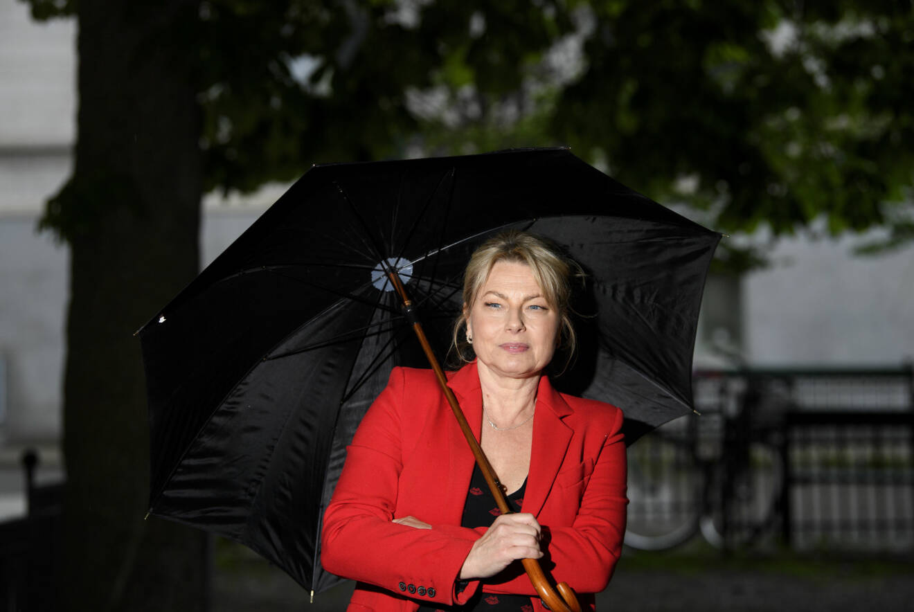 Helena Bergström håller ett paraply. Hon har en röd kavaj, blont hår i en hästsvans och blå ögon.
