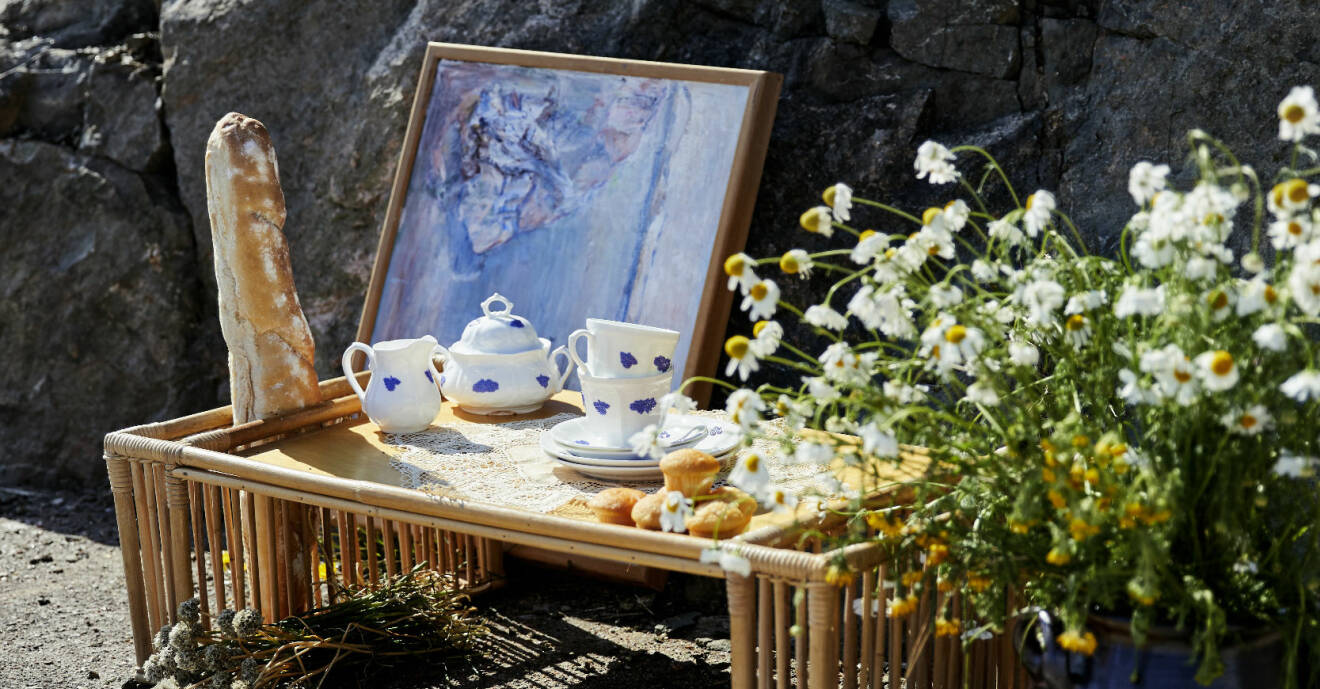 Blåvita koppar och assietter på en bänk i bambu. Bredvid står ett fång prästkragar och bakom en tavla lutad mot en stenmur.
