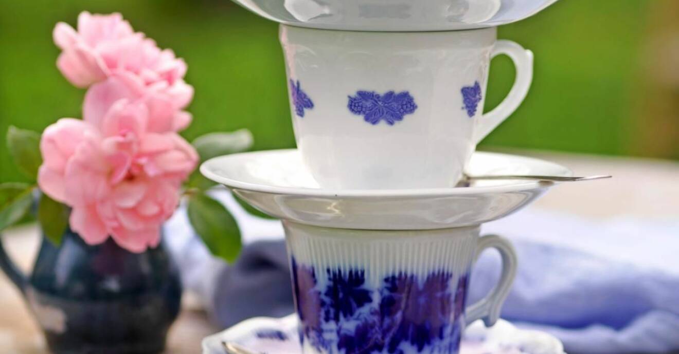 Tre blåvita kaffekoppar står staplade på varandra på ett träbord, bredvid två rosa blommor i en liten mörkblå vas.