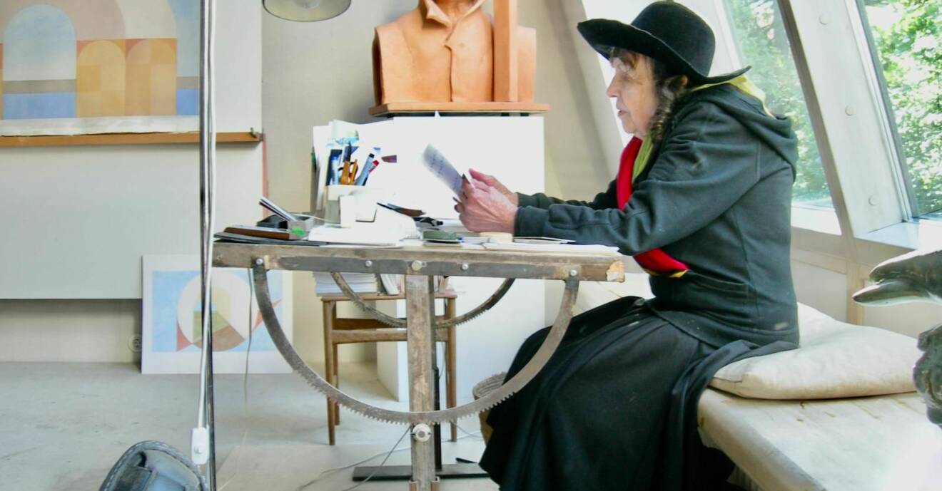 Hertha Hillfon ses från sidan, sitter vid ett bord med pennor och papper, på en träbänk och läser. Bakom henne syns en skulptur föreställande hennes man Gösta Hillfon.