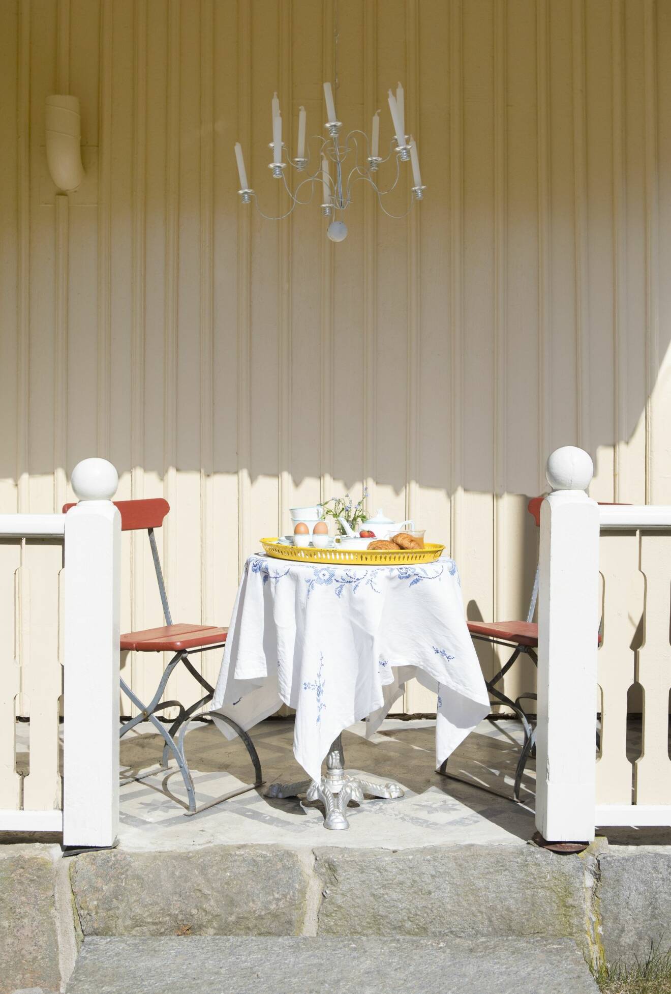 Veranda med bord och två stolar. På bordet ligger vit duk med blått mönster och en gul bricka dukad med ägg, croassianter och vit teservis.