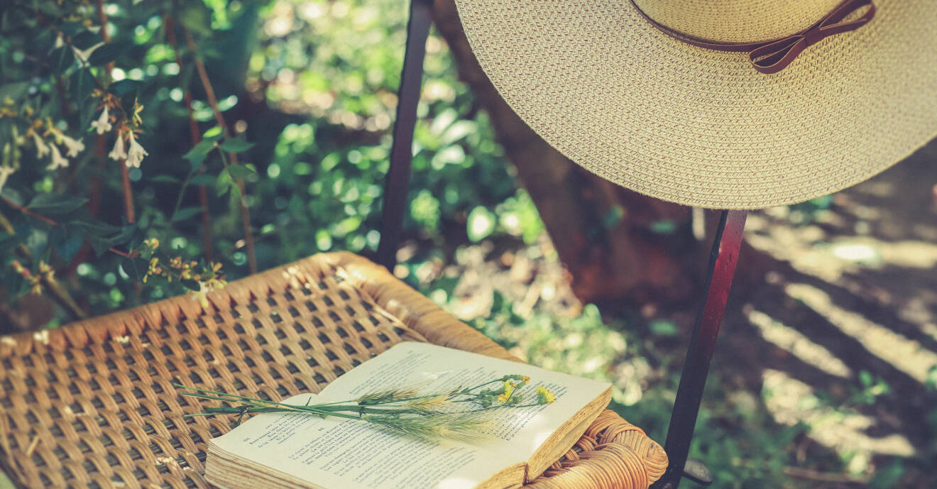 Stol med rottingsits, stråhatt hänger på stolsryggen och antik bok med blommor ligger på sitsen. Bakom syns grönska i sol.