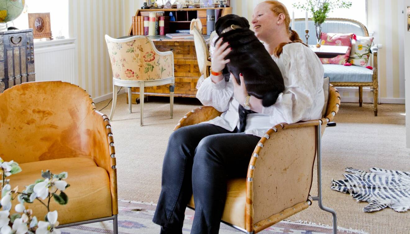 Maria Granström sitter i fåtölj med svart hund i knäet. Runtomkring syns äldre möbler och en rölakansmatta på golvet.