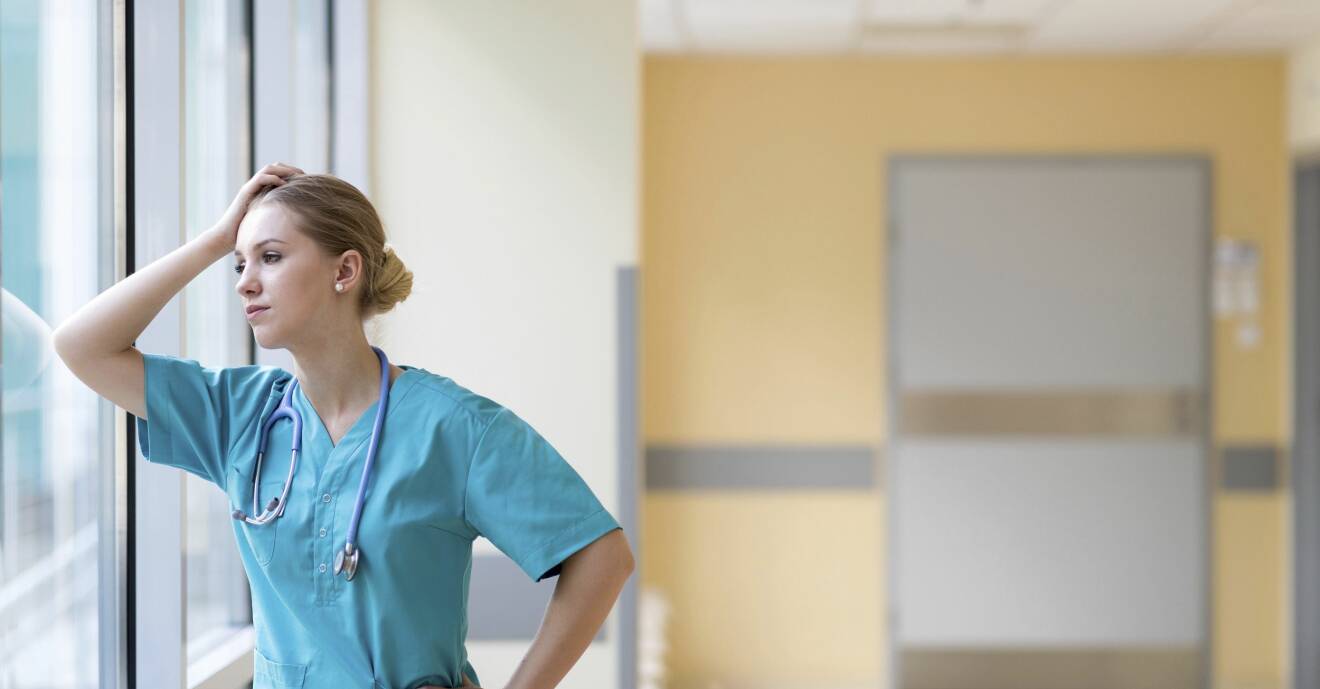 Trött sjuksköterska står lutan mot en fönsterruta