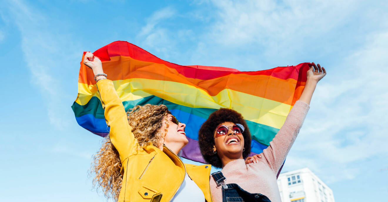 Två kvinnor ler och håller upp en Prideflagga ovanför huvudet.