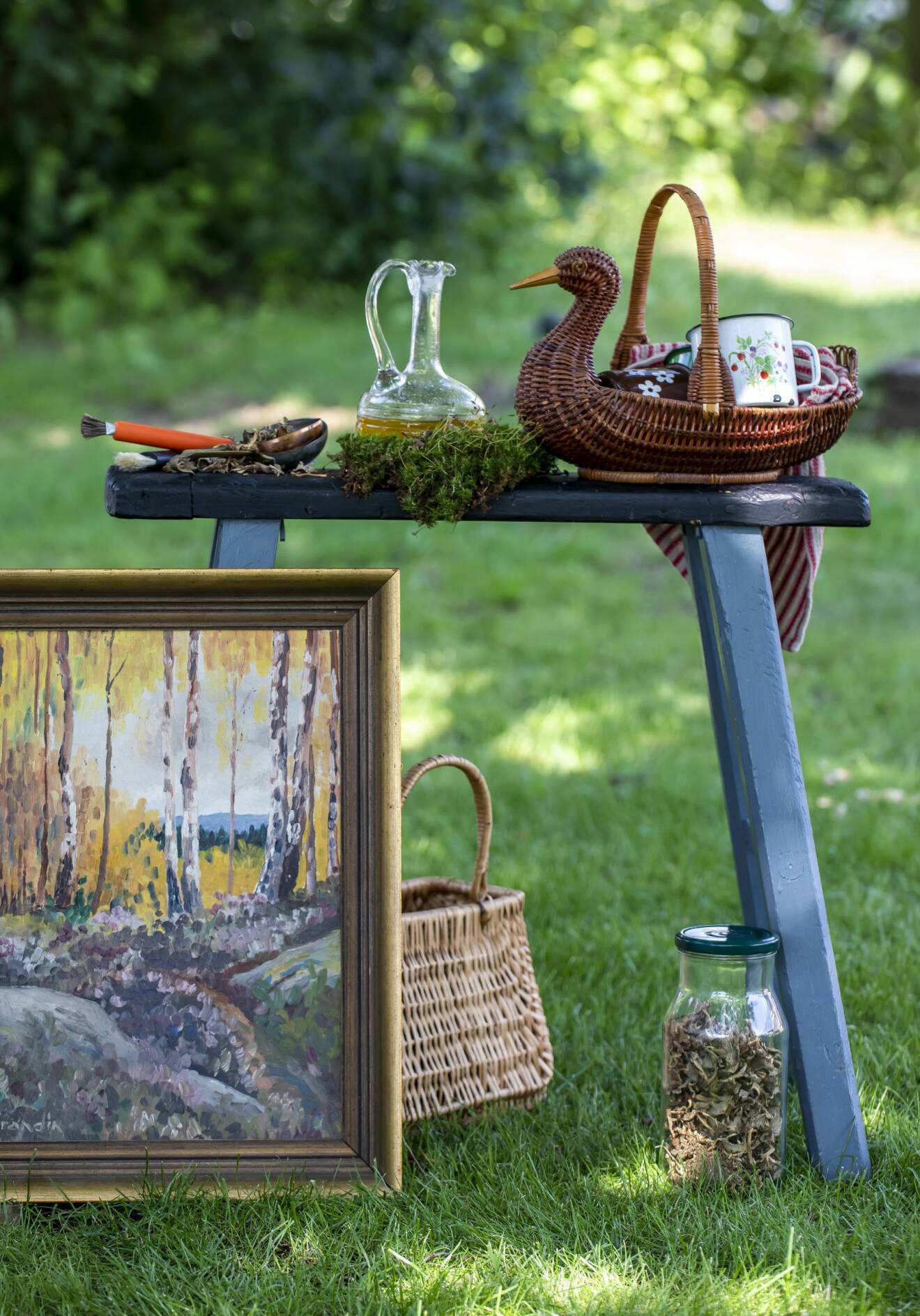 Blå allmogepall med glaskaraff och korg föreställande en and. Framför en tavla och en glasburk med svamp, grönska i bakgrunden.
