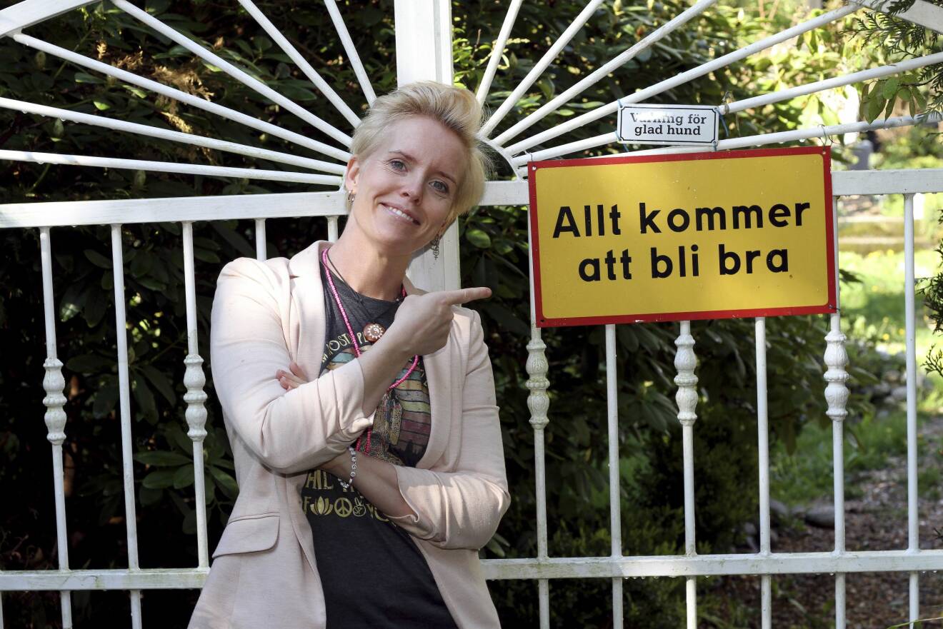 Malin Törnqvist står framför en grind. På den finns en skylt med texten "Allt kommer att bli bra". Ovanför finns en mindre skylt med texten "Varning för glad hund".