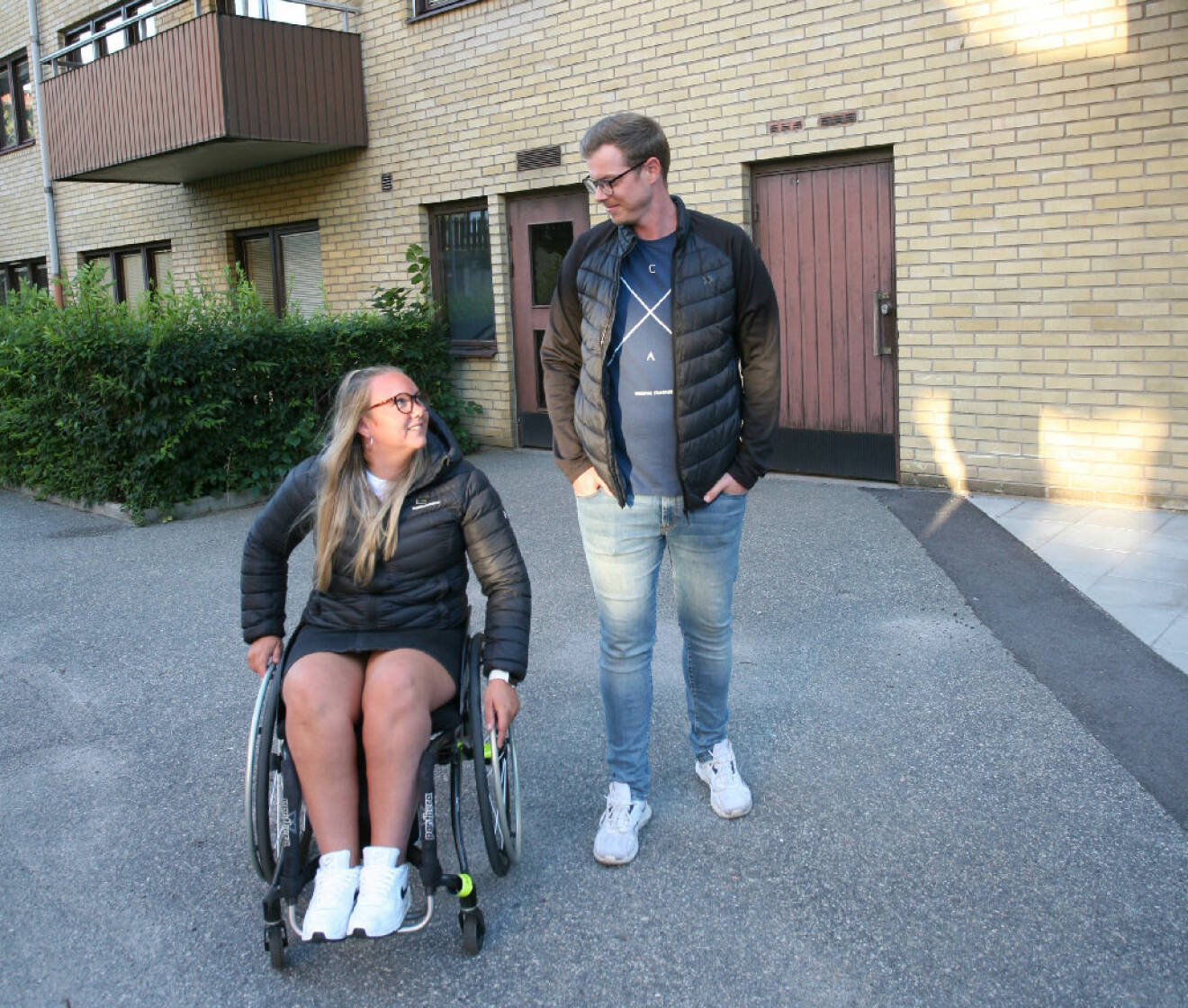 Elina Waldén i rullstol med sin Micke vid sidan om, utanför hyreshuset där de bor.