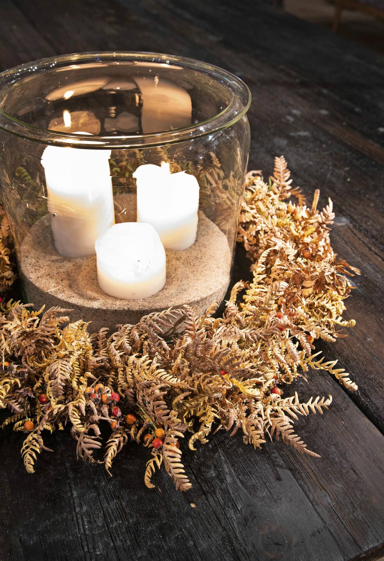 Bild på en ljuslykta i glas med tre tända stearinljus inuti. En krans av ormbunke ligger runt om ljuslyktan som står på ett brunt träbord.