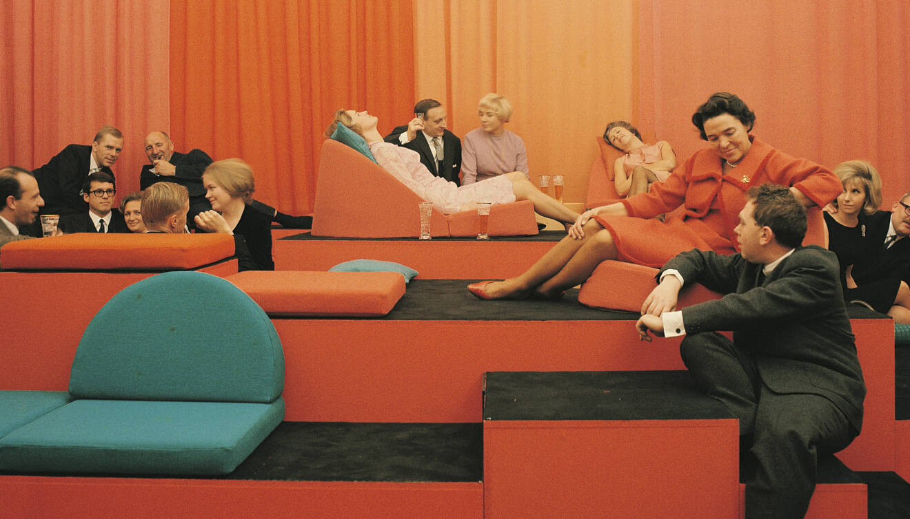 Kvinnor och män sitter i olika orange soffmoduler med orange draperi i bakgrunden.