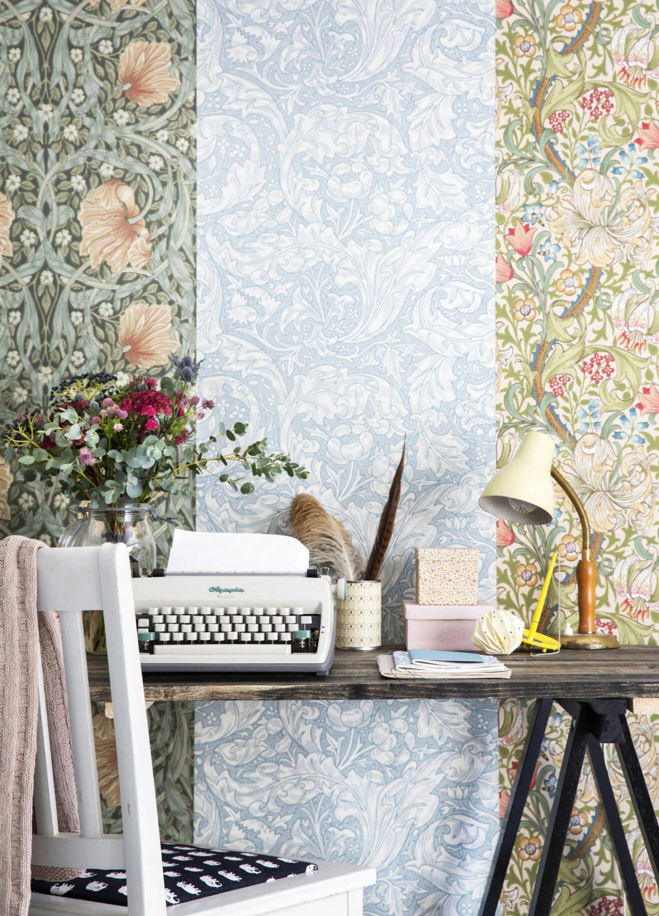 Ett skrivbord med skrivmaskin, bukett i glasvas och lampa framför vägg av tre William Morris-tapeter.