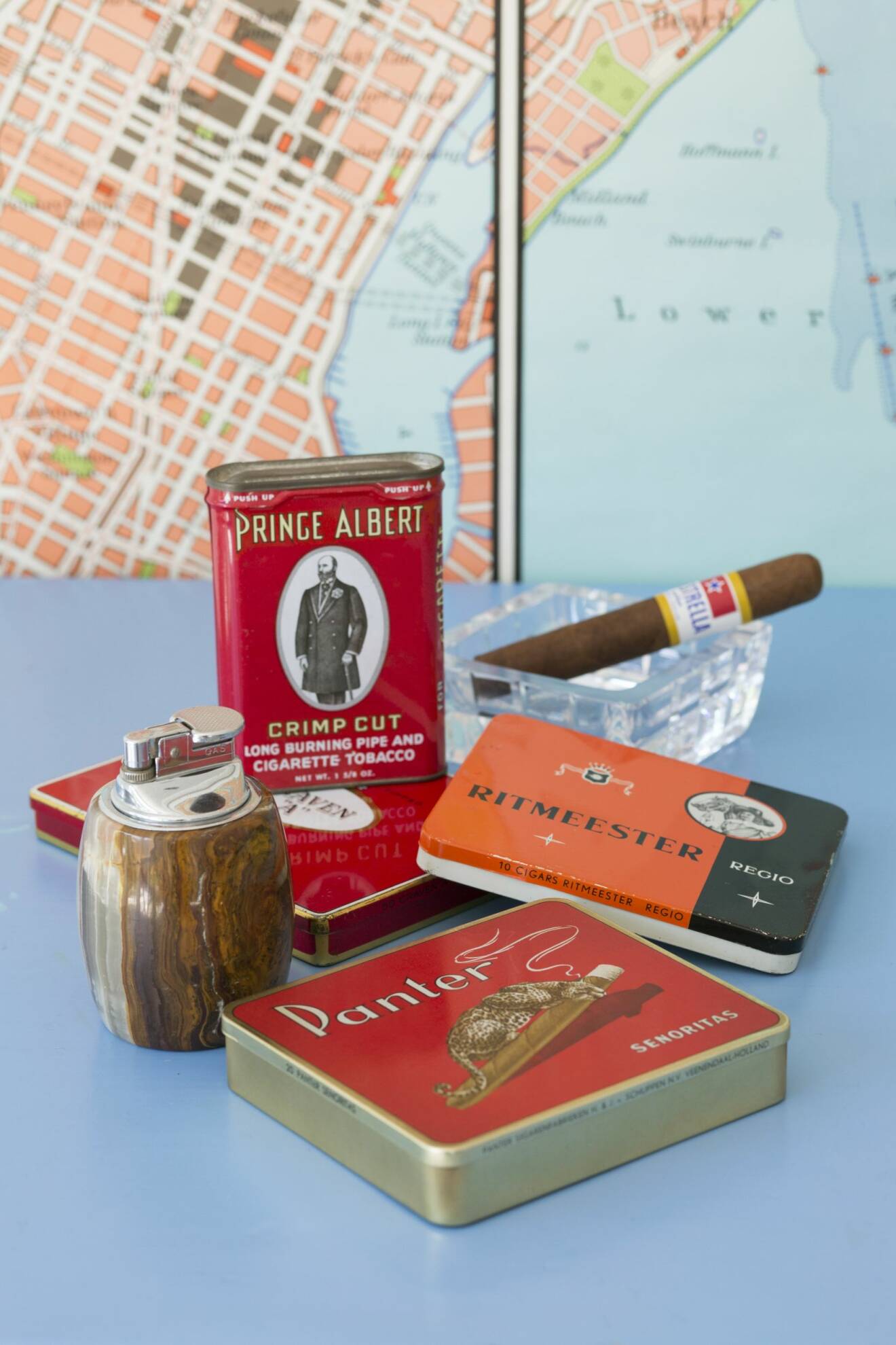 Fyra antika cigarettaskar i metall, en tändare i trä och en cigarr i ett aft av glas med karta i bakgrunden.