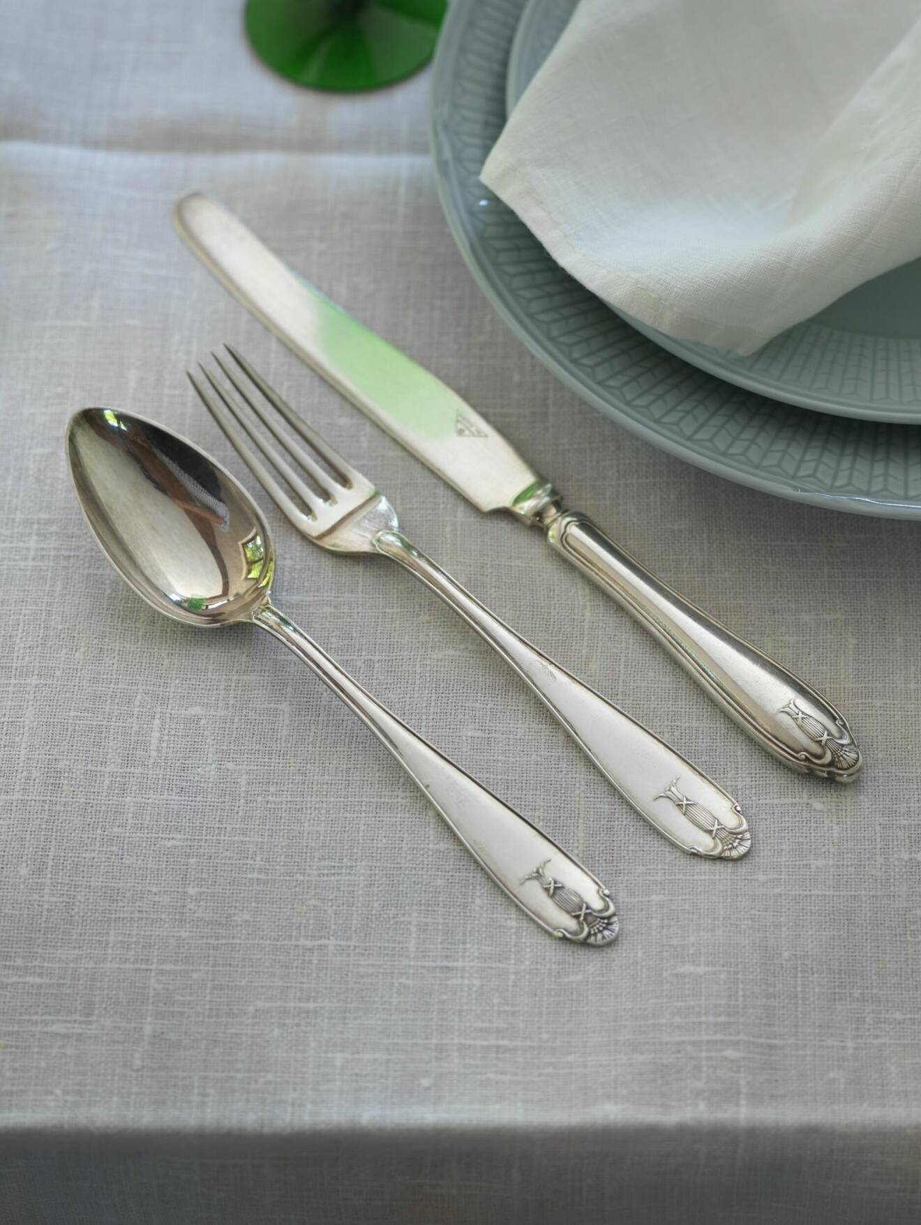 Sked, gaffel och kniv på grå dul. Två grå swedish grace-tallrikar med vit servett på syns till hälften.