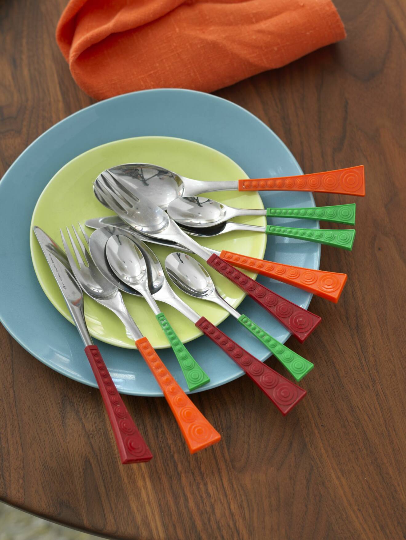 Gafflar, knivar och skedae med grönt, vinrött och orange skaft på ljusgrön och ljusblå tallrik, orange servett bredvid på mörkt träbord.
