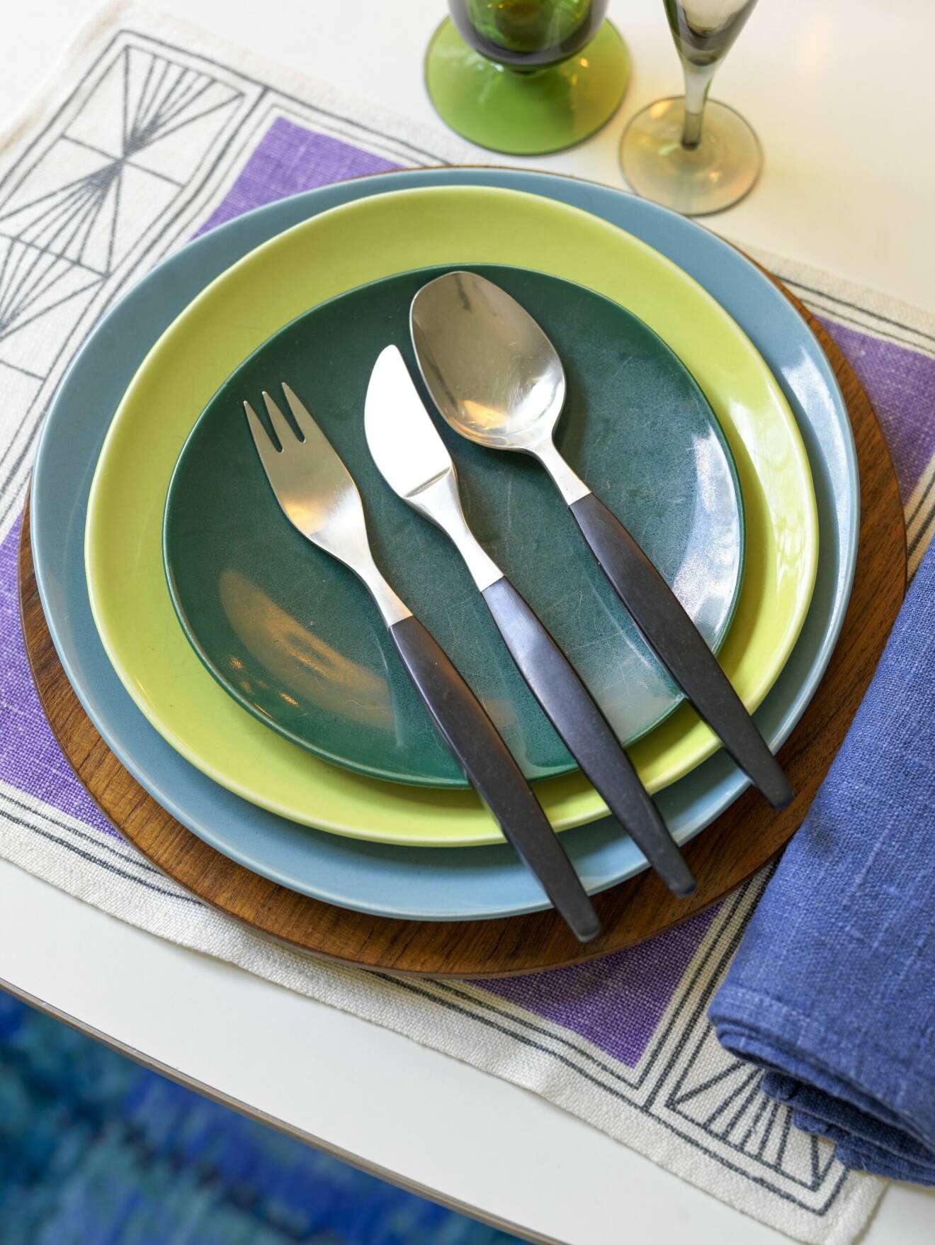 Tre tallrikar i ljusblått, limegrnt och mörkgrönt staplade, gaffel, kniv och sked med svarta handtag ligger på tallrikarna.
