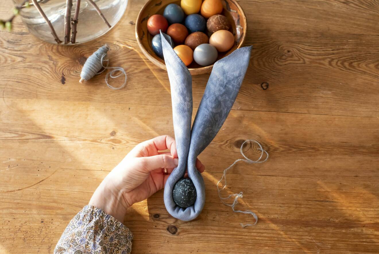 Träskål med färgade ägg och en blå tygbit som vikts och formats som kaninöron runt ett färgat ägg.
