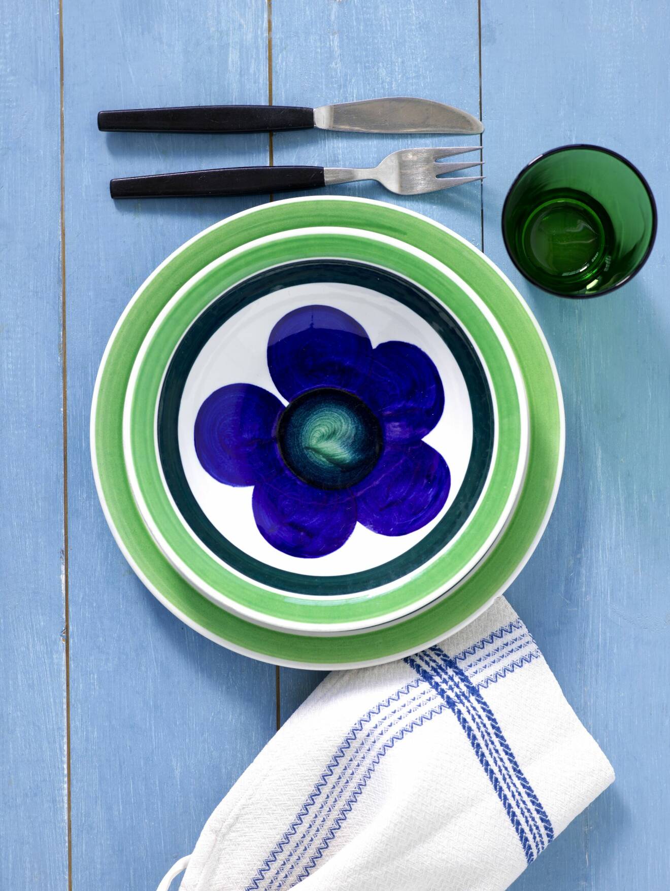 Tallrikar från Marianne Westmans serie Sippa i grönt, vitt och blått med gaffel och kniv med svart skaft och ett grönt glas på ett blått träbord.