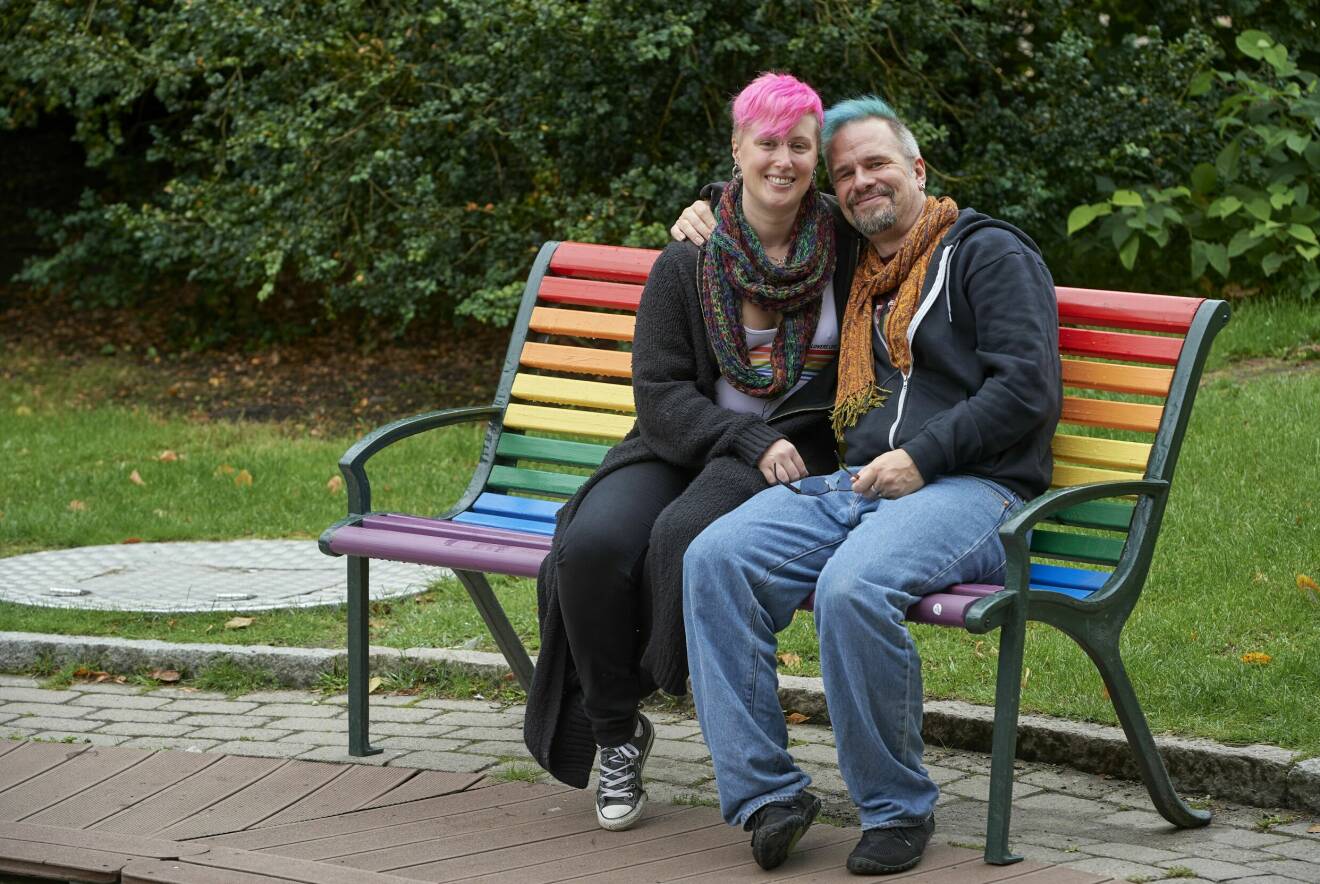 Polyamorösa paret Anna och Patrik sitter på en parksoffa som är målad i regnbådens färger.