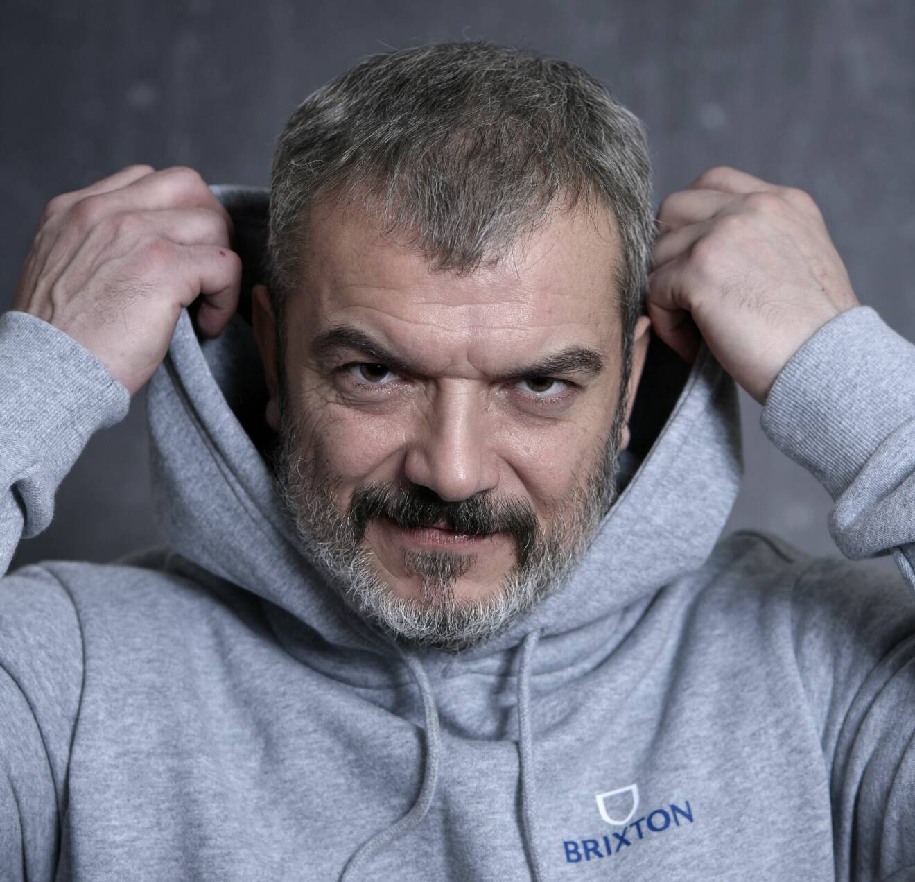 Skådespelaren Dragomir Mrsic drar upp huvan på sin grå hoodie.