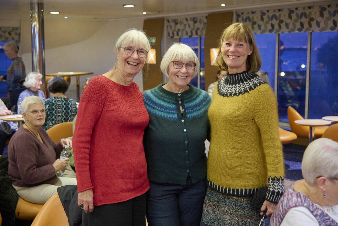 Tre glada damer har rest sig upp från borden och poserar i sina hemstickade tröjor.