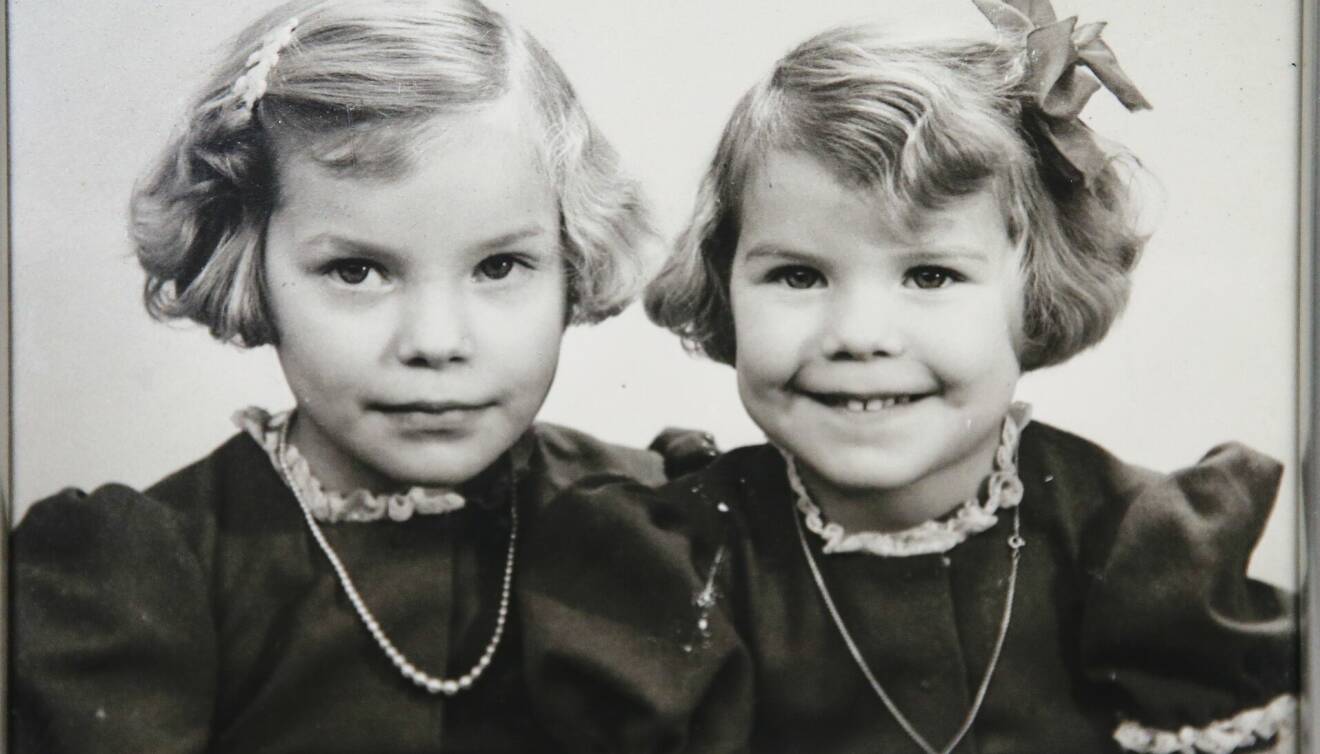 Systrarna Marianne och Ann-Katrin är finklädda i klänning med puffärm och rosett i håret och sitter bredvid varandra.