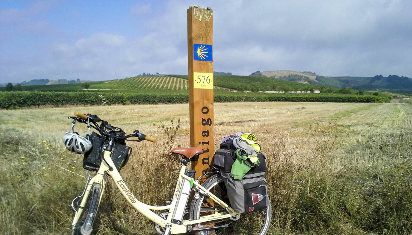 Jeannets cykel står lutad mot en träpåle med angivelsen 576 kilometer kvar till Santiago Compostela och i bakrgunden syns fält, bland annat en vinodling.