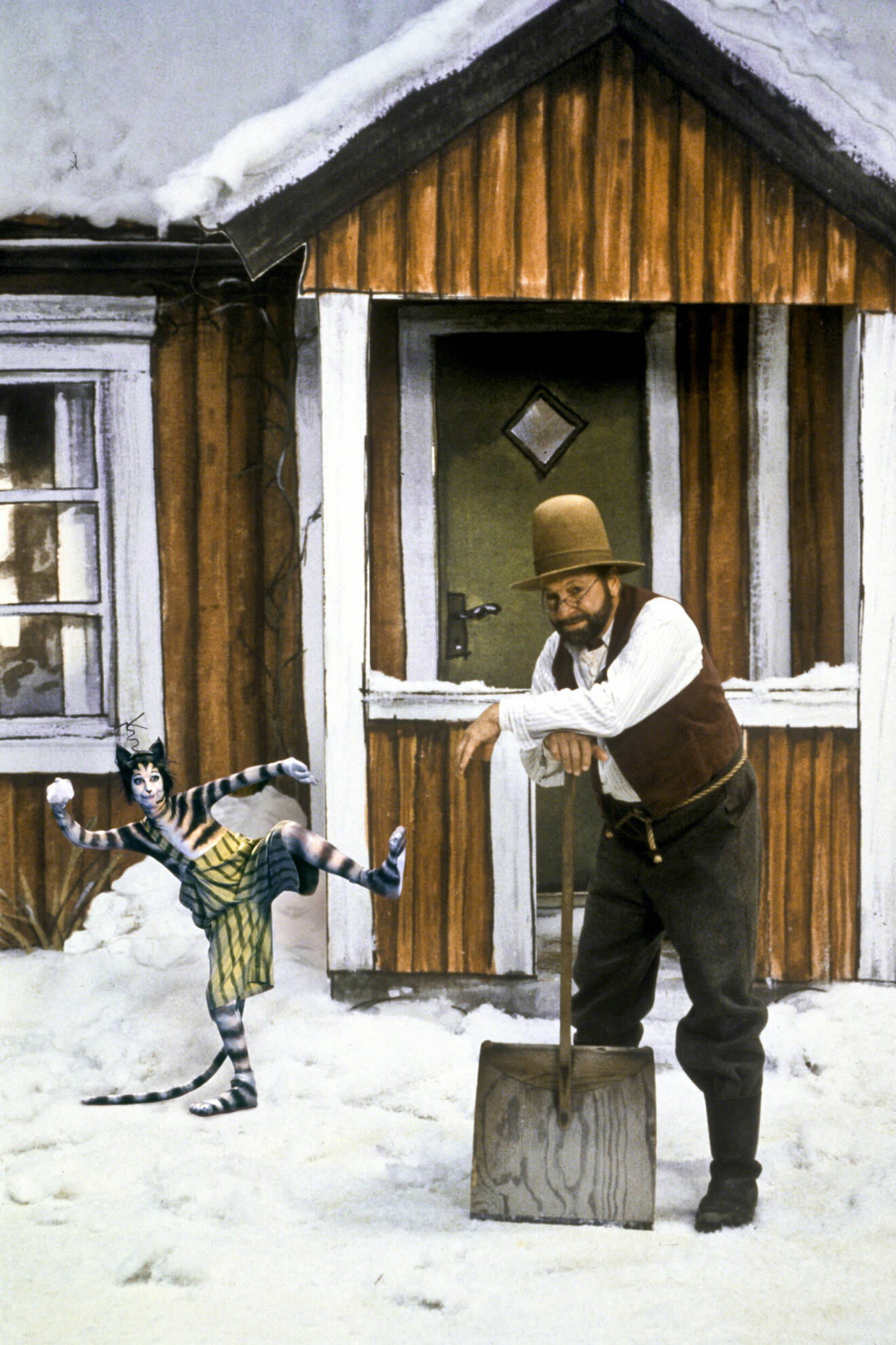 Bild på Pettson och Findus som står framför ett rött trähus i julkalendern Tomtemaskinen. Det ligger snö på taket och på marken. Findus håller i en snöboll och Pettson lutar sig på en snöskyffel.