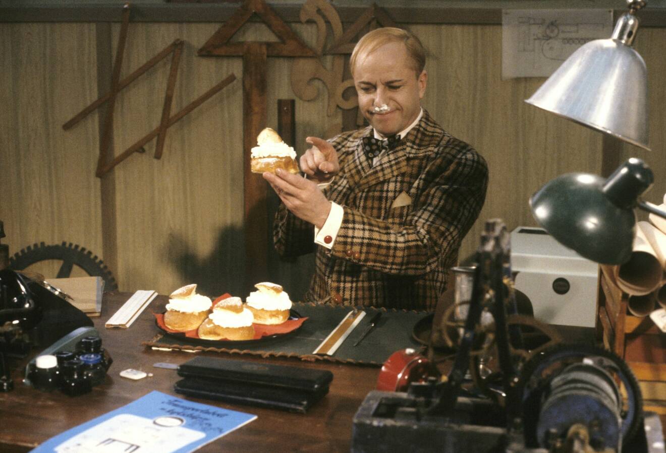 Bild på skådespelaren Helge Skoog som karaktären Ture Sventon i julkalendern. Han sitter i ett kontor och har ett fat med semlor på bordet framför sig. Han håller en semla i handen och har grädde på näsan.