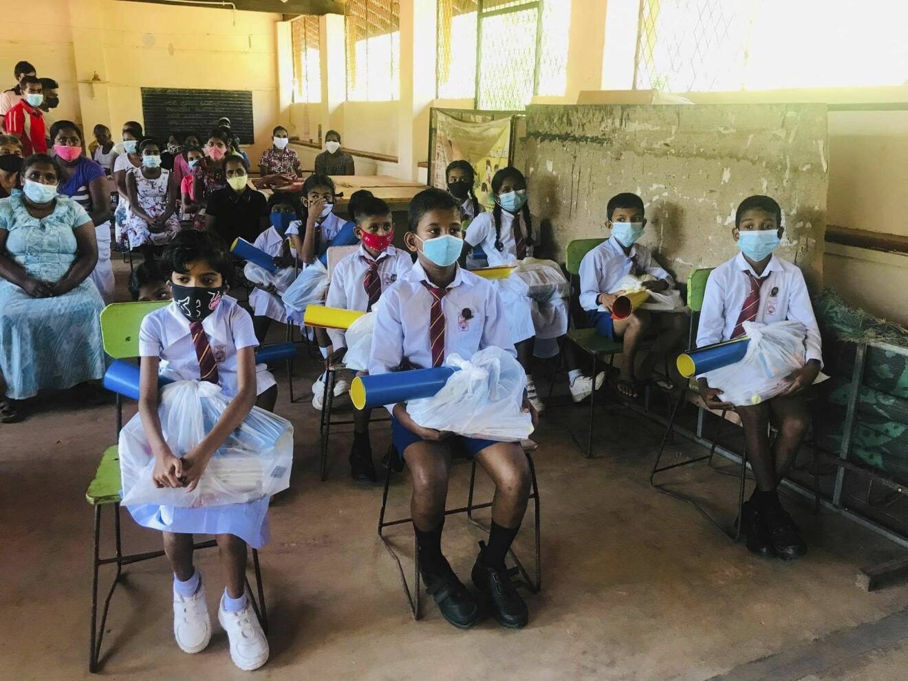 Barnen på en skola i Sri Lanka sitter med gåvopåsar i knät. Alla bär munskydd.